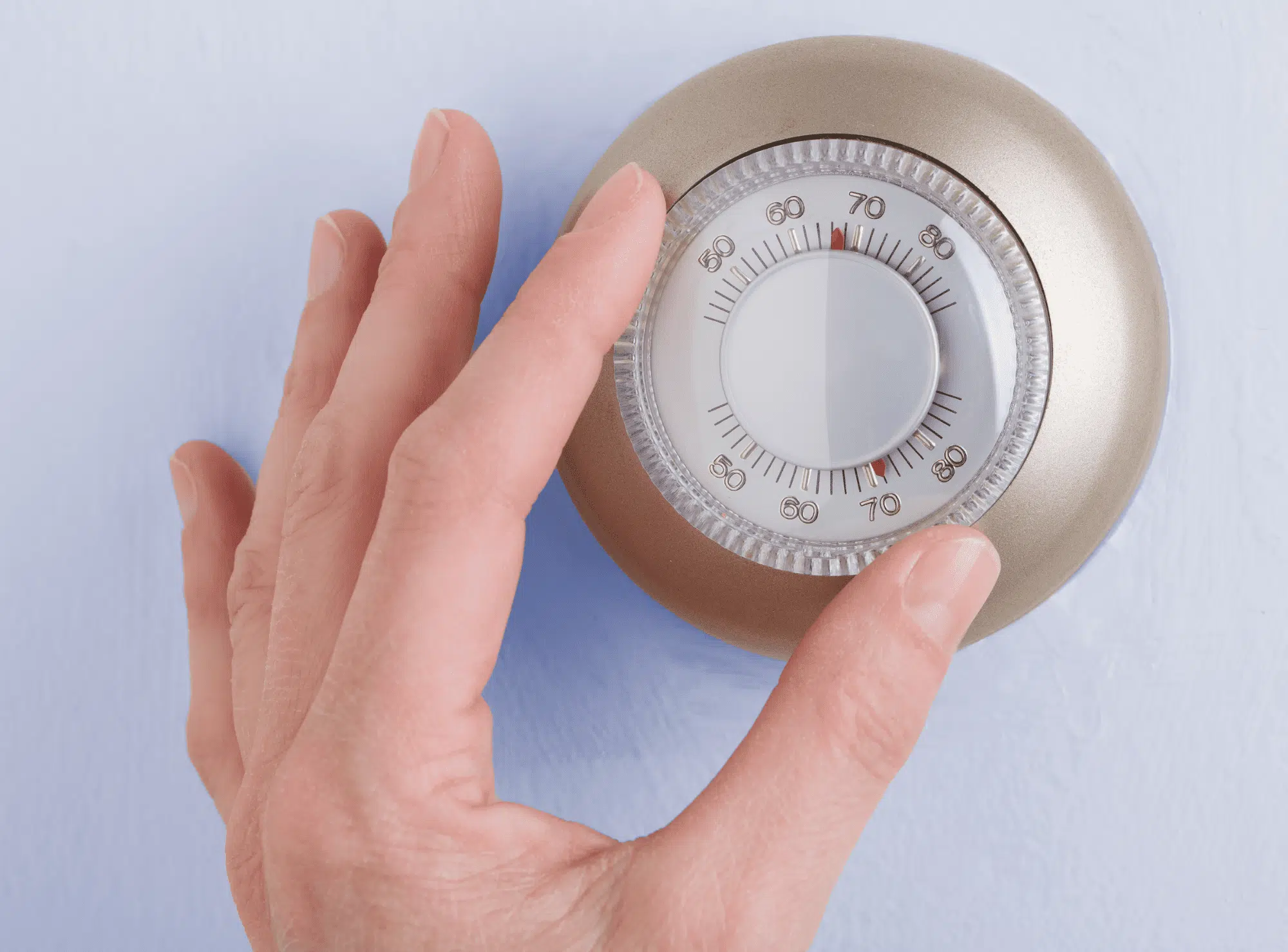 A hand adjusting a classic round mechanical thermostat on a light blue wall during a temperature check from Right Time HVAC.