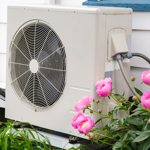 A white outdoor HVAC condenser unit mounted on the exterior wall of a house, positioned near blooming pink peonies in a garden setting.