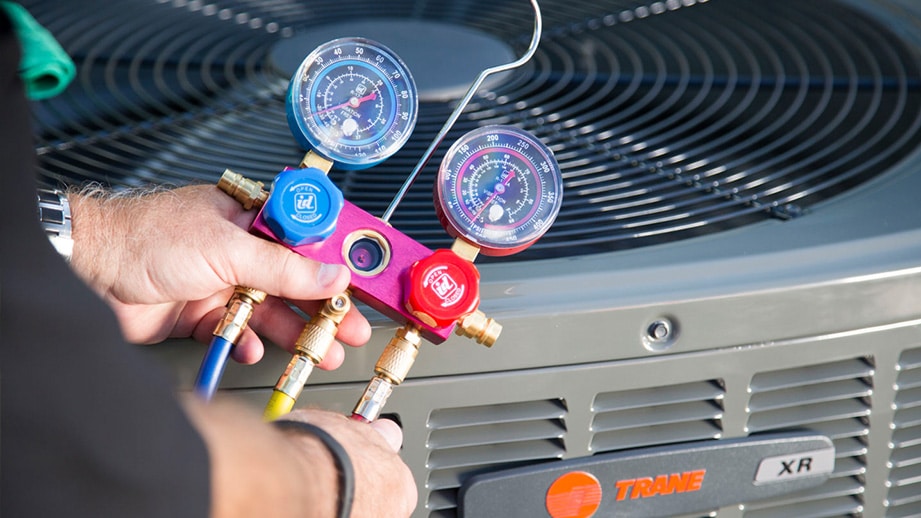 Spring AC Tune Up: A technician's hands holding a manifold gauge set with blue and red dials over a Trane XR air conditioning unit to check refrigerant pressure.