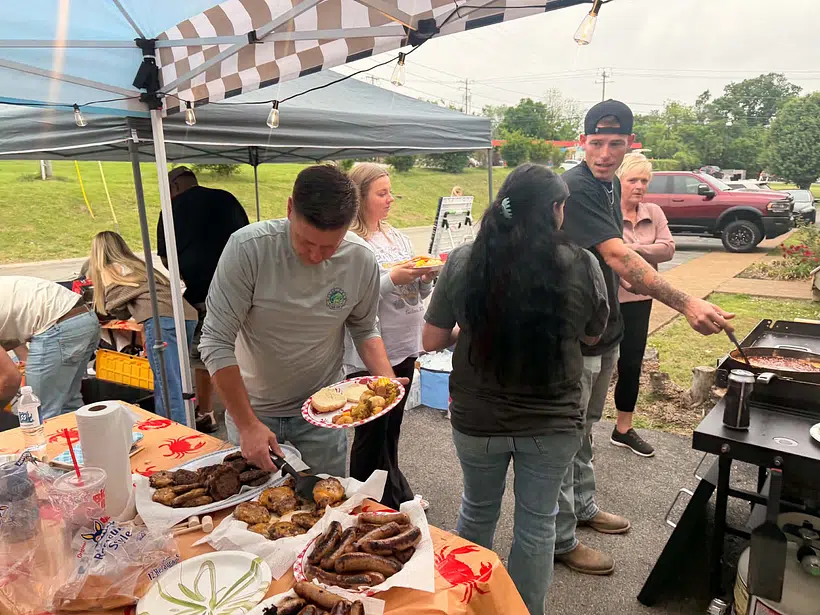 An outdoor company cookout under canopies where team members of Right Time HVAC serve food, grill on a flat-top, and chat around a table piled with burgers and sausages.